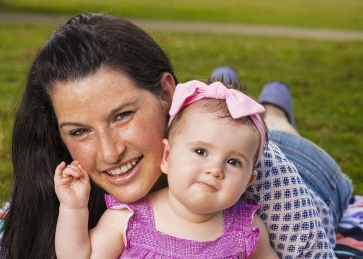 A woman with long dark hair lies on a picnic rug holding a young baby dressed in a pink dress and headband.