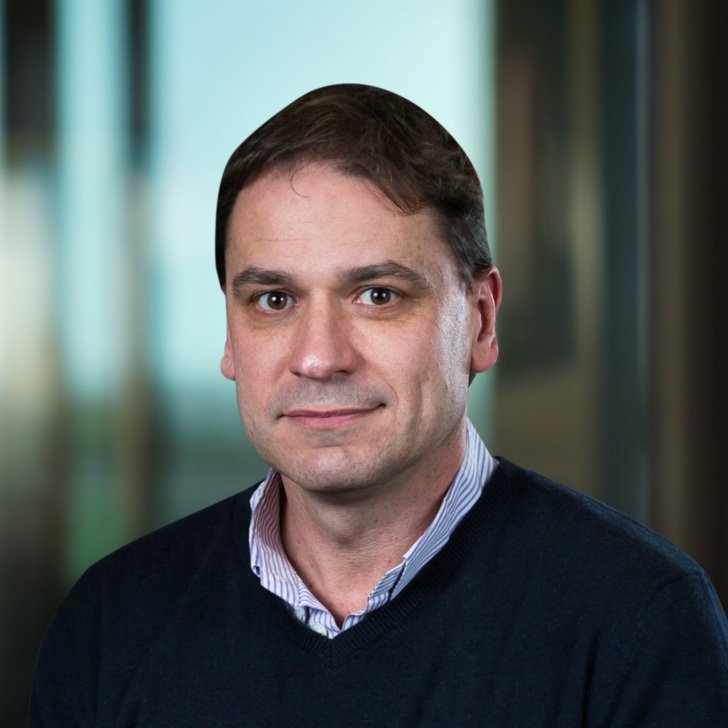 Man with dark hair wearing a light blue collared shirt and a dark blue jumper