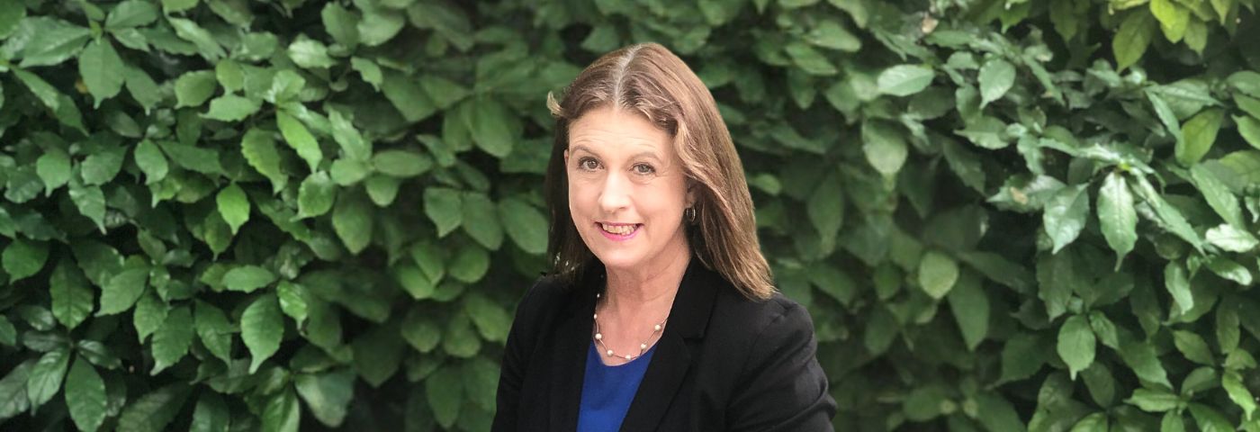 Female research with shoulder-length brown hair wearing a black jacket and standing in front of green foliage.