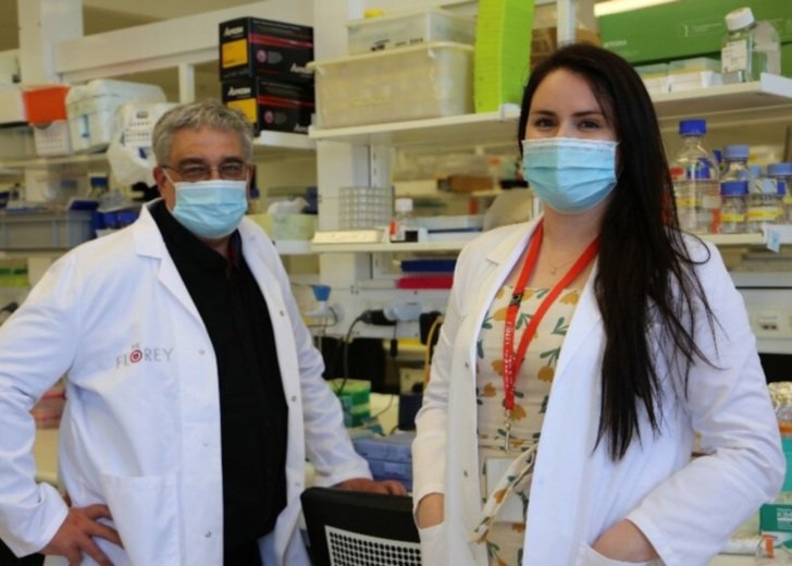 One male and one female researcher wearing white lab coats and blue face masks and standing in a Florey laboratory.