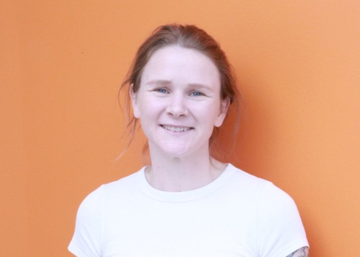 Female researcher standing against a pale orange backgound. She has light brown hair pulled back and wears a white tshirt.