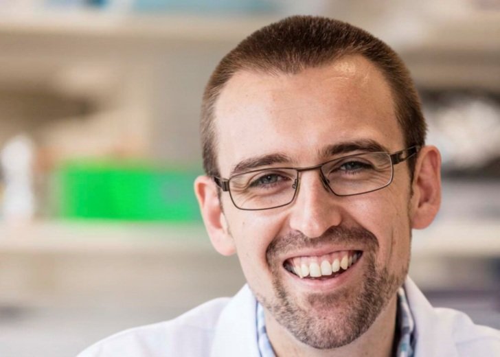 Male researcher with short brown hair and glasses. He wears a checked shirt with a white lab coat.