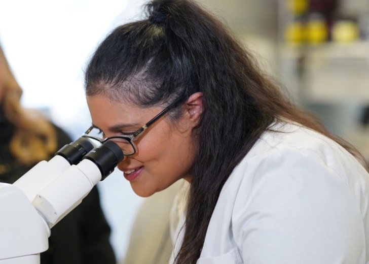 A woman with dark hair and glasses and wearing a white lab coat and looks into a microscope.