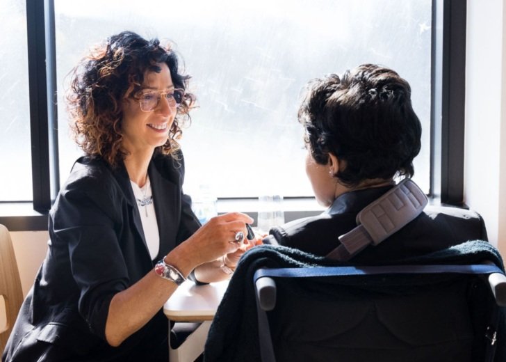 Woman with curly hair and black jacket sitting with a young woman in a wheelchair who has had a stroke.