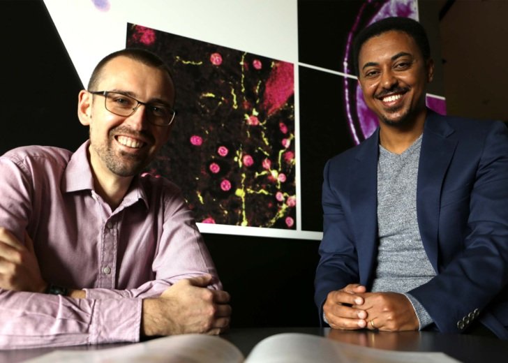 Two men sit pictured in front of microscopic images