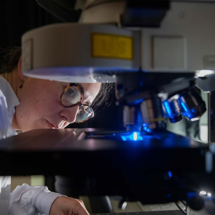 Researcher wearing lab coat peers into microscope in dark room