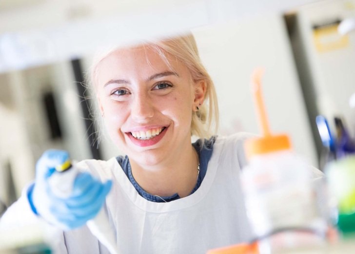 Young female researcher looks up at camera and smiles while pipetting in a lab