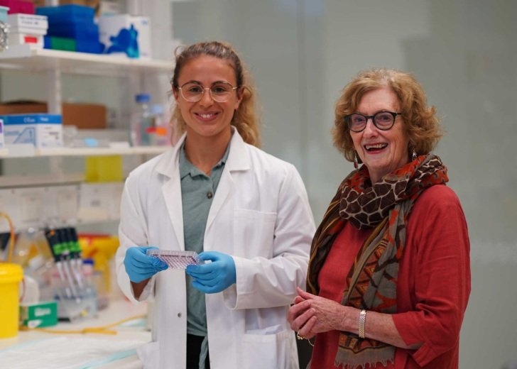 Young female researcher in white lab coat and blue gloves standing next to a female donor in a Florey laboratory.