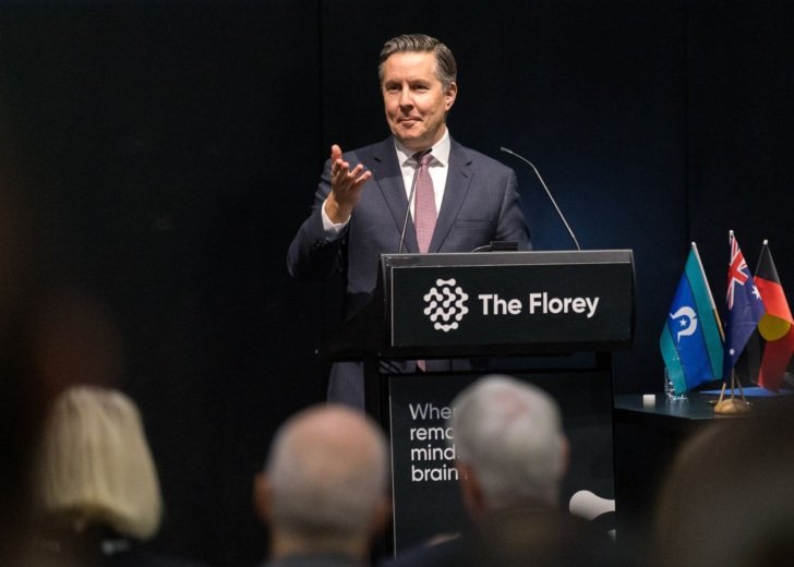 The Hon Mark Butler MP standing at a podium with a placard reading 'The Florey'. He is wearing a suit and tie and looking to the audience with one of his hands raised.