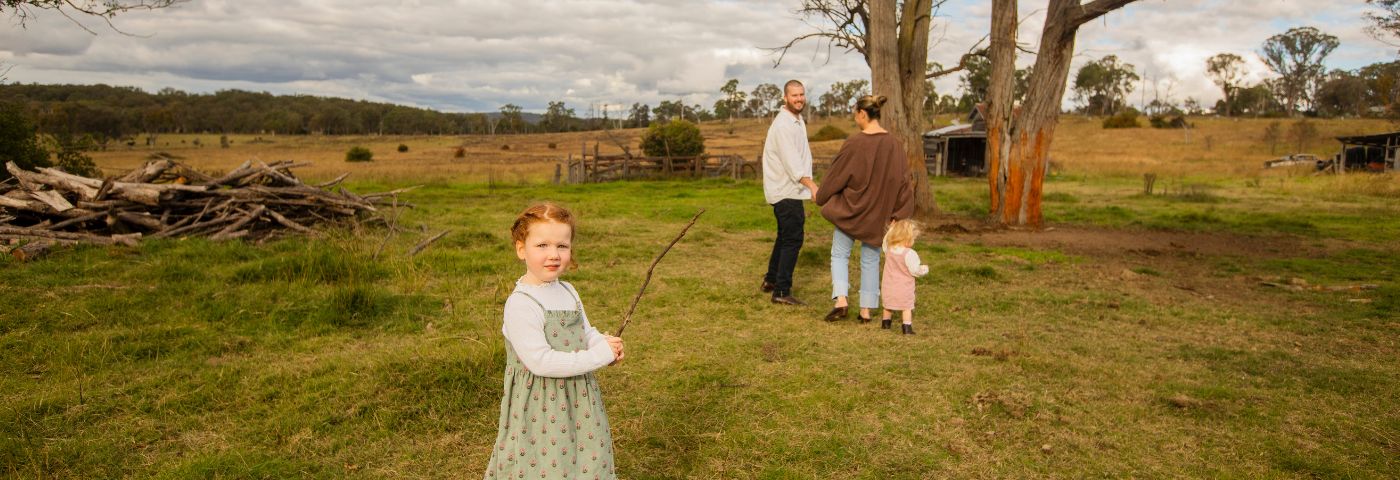 Young girl with red hair and a green dress holding a stick. She is on a rural property with bushland in the background.