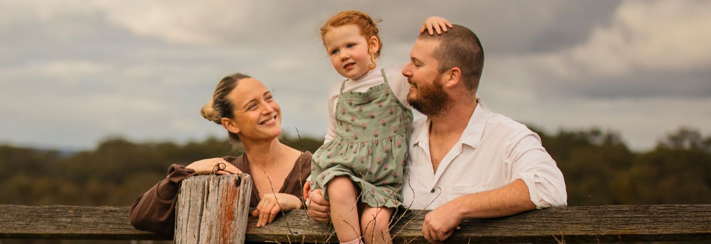 A young girl with red hair and a green dress sits on an old wooden fence between her mother and father.