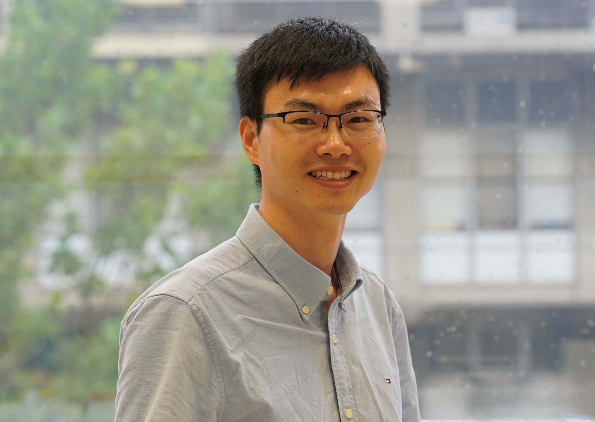 Dr Huan Liao standing against a window in a Florey laboratory. He wears a blue collared shirt and glasses.
