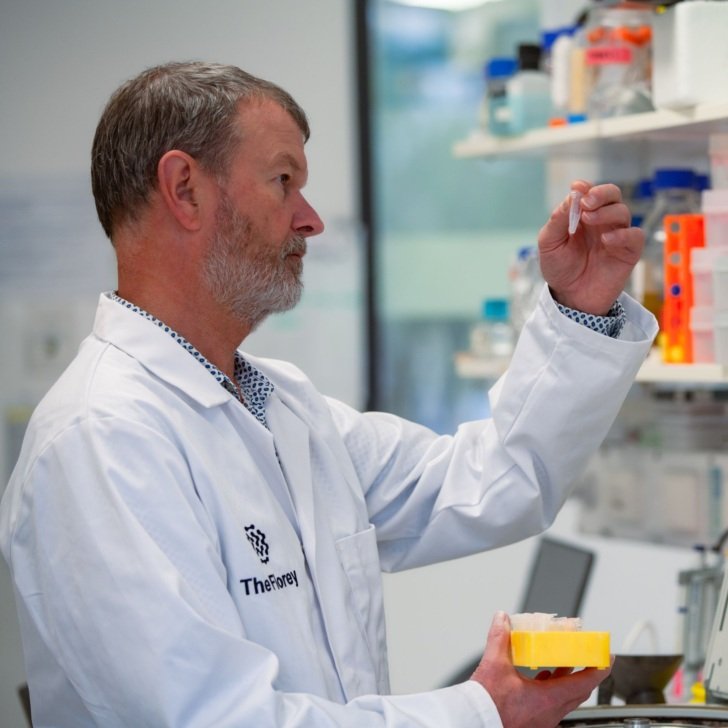 Male researcher with white Florey laboratory coat holding up and inspecting solution in a small tube.