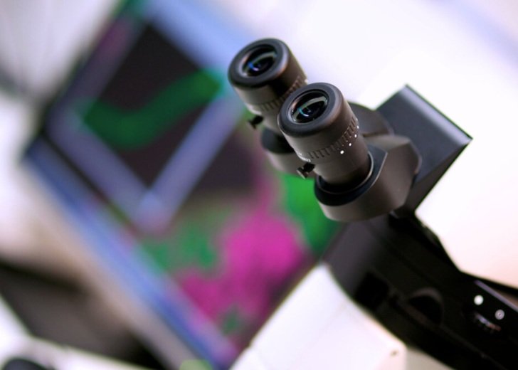 Close up of a microscope, showing the eyepiece and blurred imagery on a computer screen in the background.