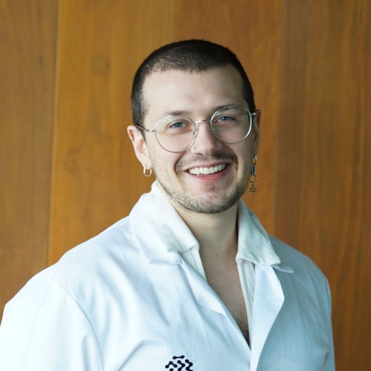 Jarrah Kron, PhD student at The Florey. They are wearing a white lab coat and standing against a brown wood wall.