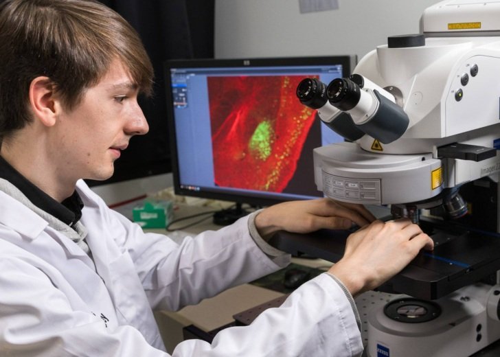 Young male researcher works at a Florey microscope. A computer screen in the background shows a vivid red microscopy image.