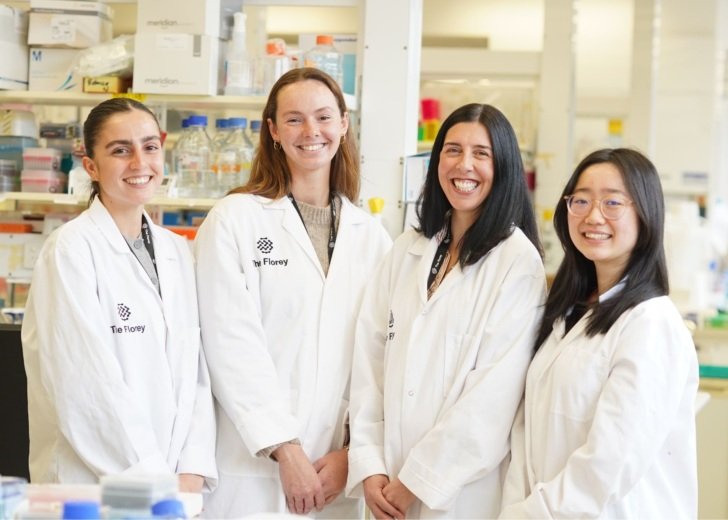 Four women researchers standing in a Florey laboratory wearing white lab coats and smiling.