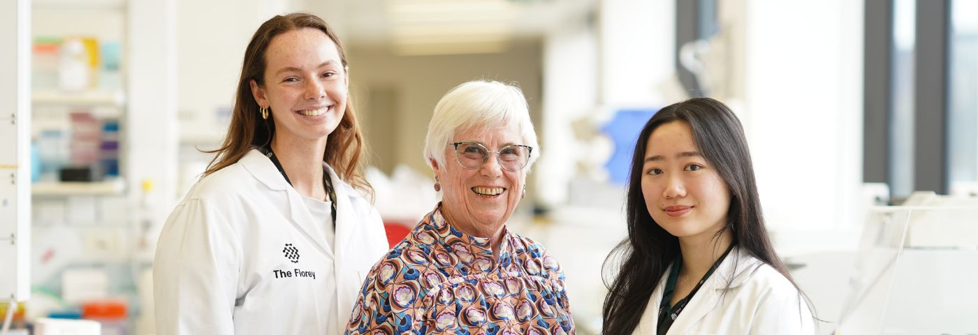 An older woman with grey hair and glasses standing between two young women researchers in white lab coats.