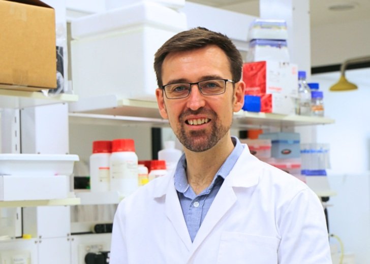 A man with brown hair and glasses wearing a blue shirt and white laboratory coat.