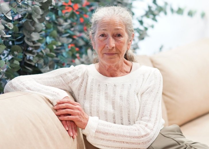 Older woman with grey hair tied back and wearing a white jumper sits on a beige couch.