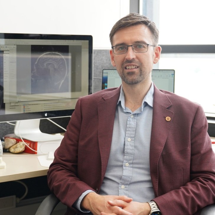 Man with brown hair and glasses wearing a jacket and collared shirt and sitting on a chair in an office. Two computer screens are in the background.