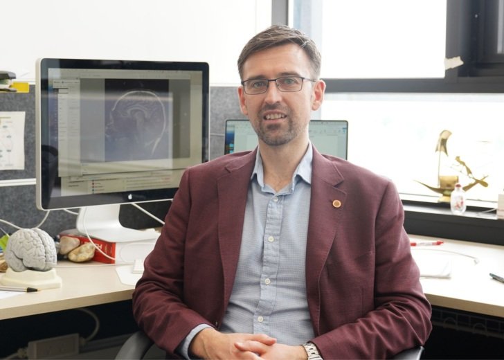 Man with brown hair and glasses wearing a jacket and collared shirt and sitting on a chair in an office. Two computer screens are in the background.