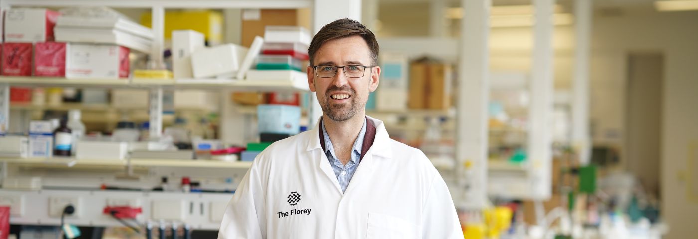 Man with brown hair and glasses wears a white laboratory coat and stands in a Florey lab in front of a bench, with shelving behind.