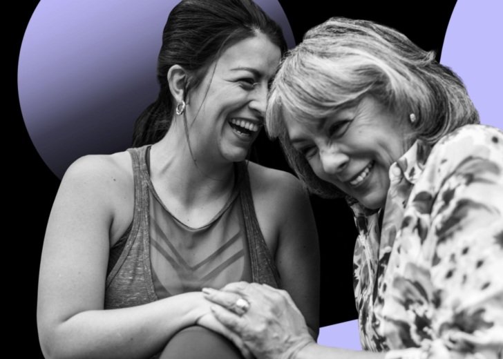 Two women holding hands and laughing. One is younger and wears active wear, the other is older and wears a patterned shirt.