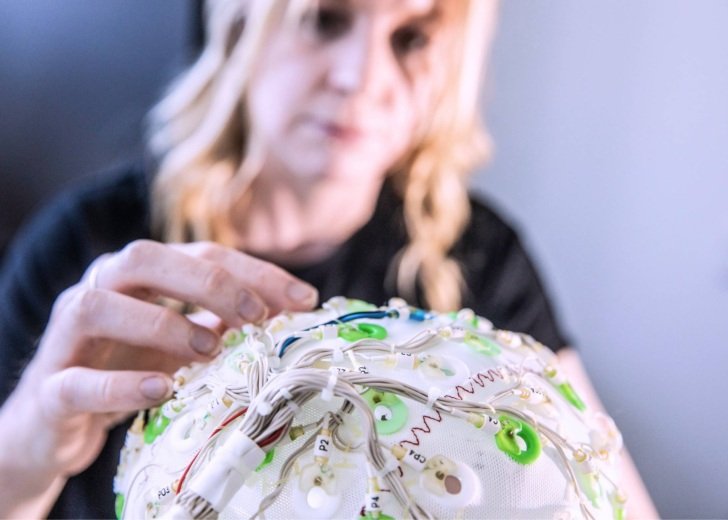 Woman with blonde hair and black shirt holds an EEG cap.