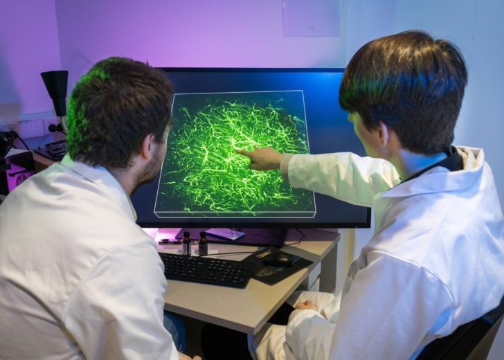 Two men wearing white laboratory coats seated at a desk and looking at a computer screen with a bright green scientific image