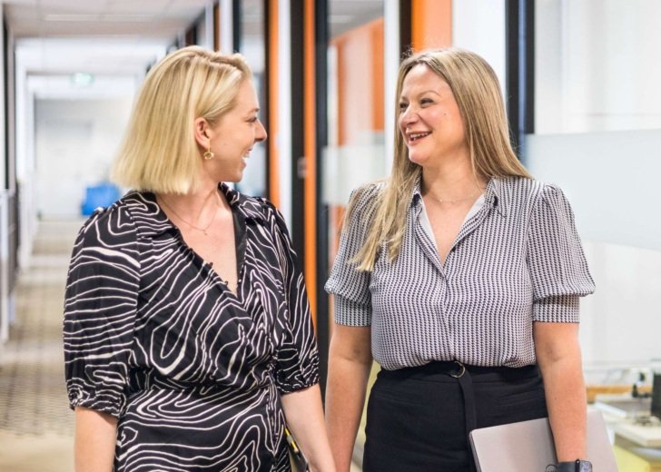 Two women talking and smiling while walking down a corridor in an office building