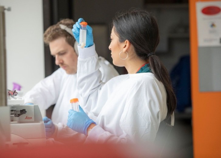 A woman with dark hair holds up a test tube with solution in her right hand. She looks at it closely while in the background a male researcher can be seen working.