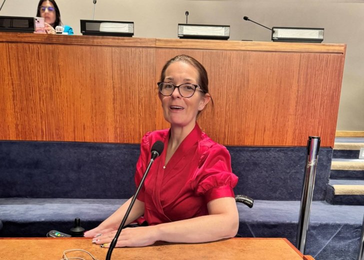 Woman wearing glasses and a pink shirt sits at a table with a microphone in front of her