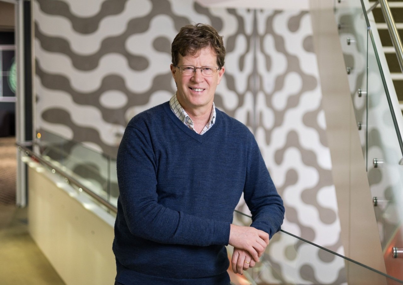 Man wearing navy jumper and smiling stands in open corridor