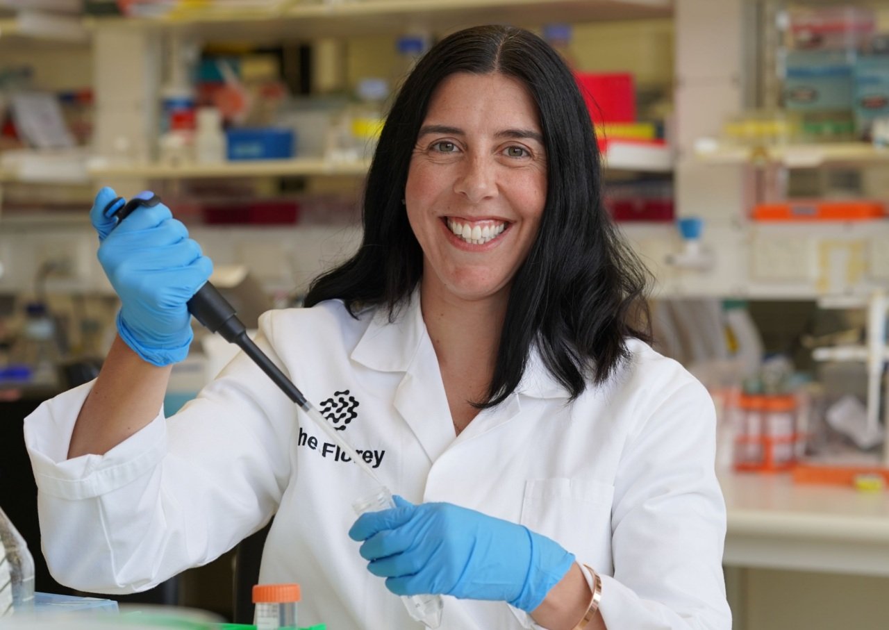 Scientist smiles wearing lab coat and holding pipette.