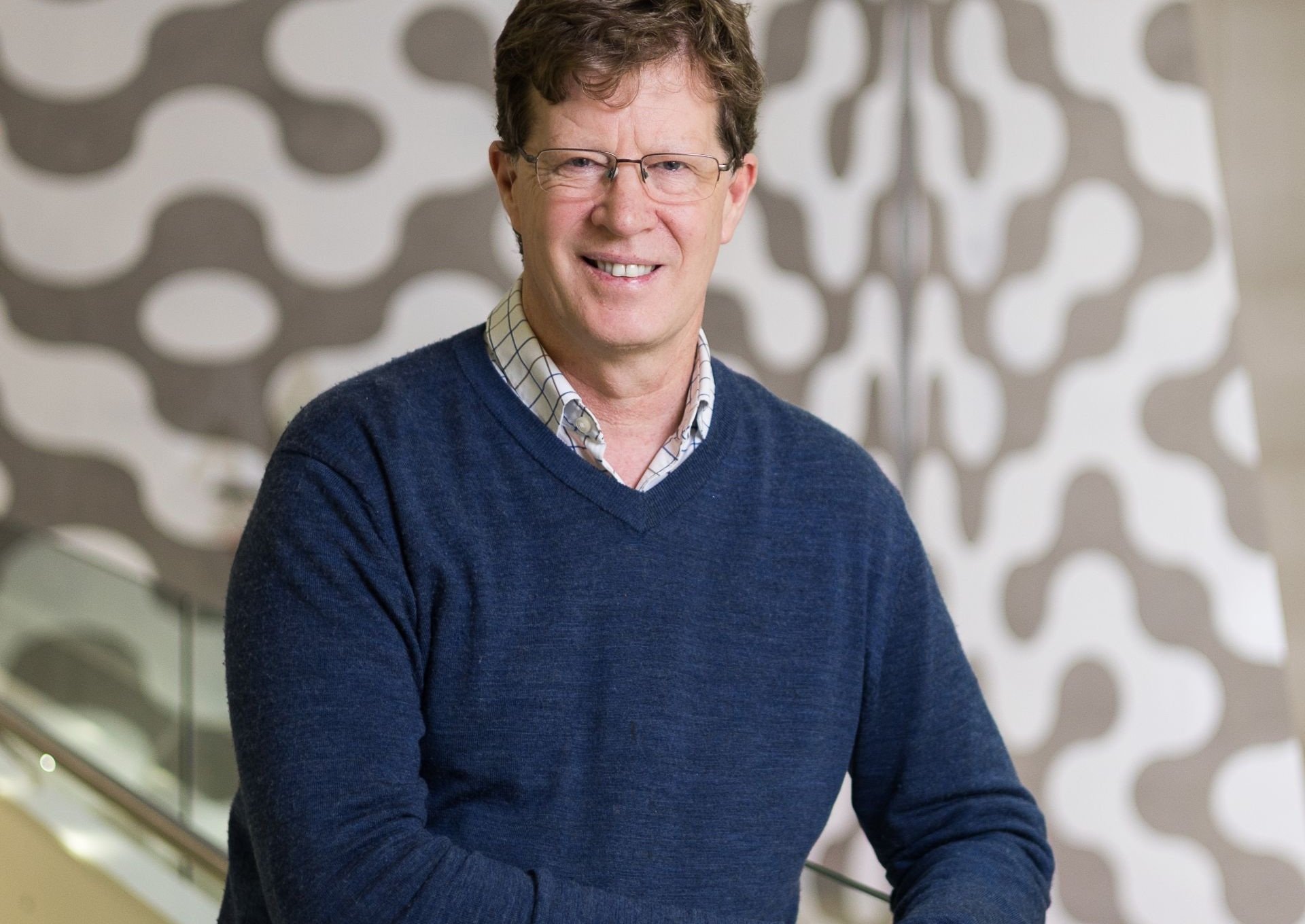 Man in navy jumper stands in open corridor, smiling