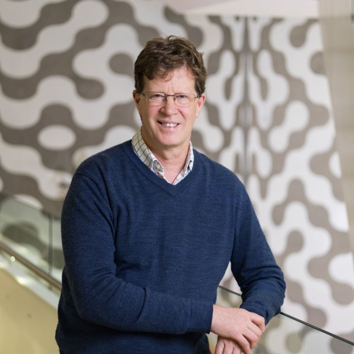 Man in navy jumper stands in open corridor, smiling