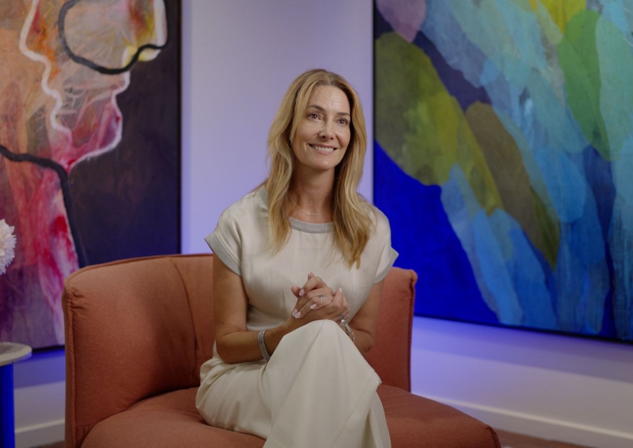 Woman sits on couch smiling with colourful artwork behind her