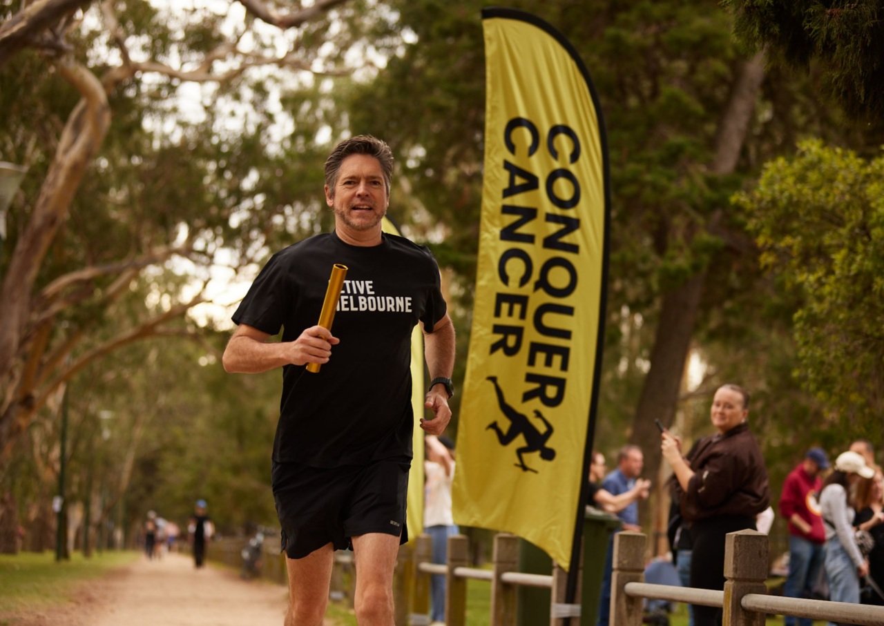 Man jogging around park next to Conquer Cancer sign holds gold baton