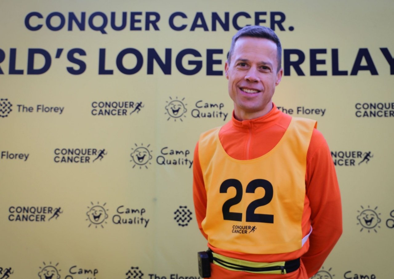 Man wearing yellow running bib smiles in front of Conquer Cancer media wall