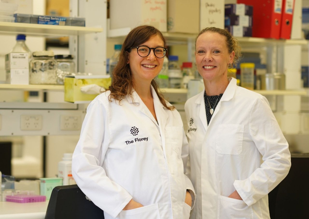 Two scientist wearing white lab coats stand smiling